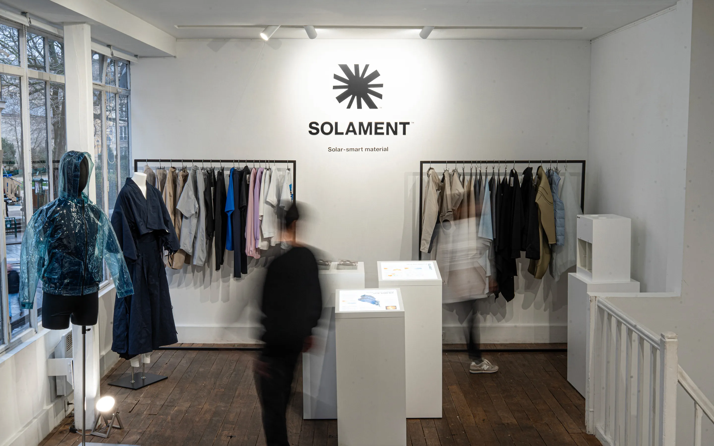 A visual of an exhibition in Paris, with various garments—including a blue transparent jacket—displayed beneath the SOLAMENT™ wall logo as visitors are around