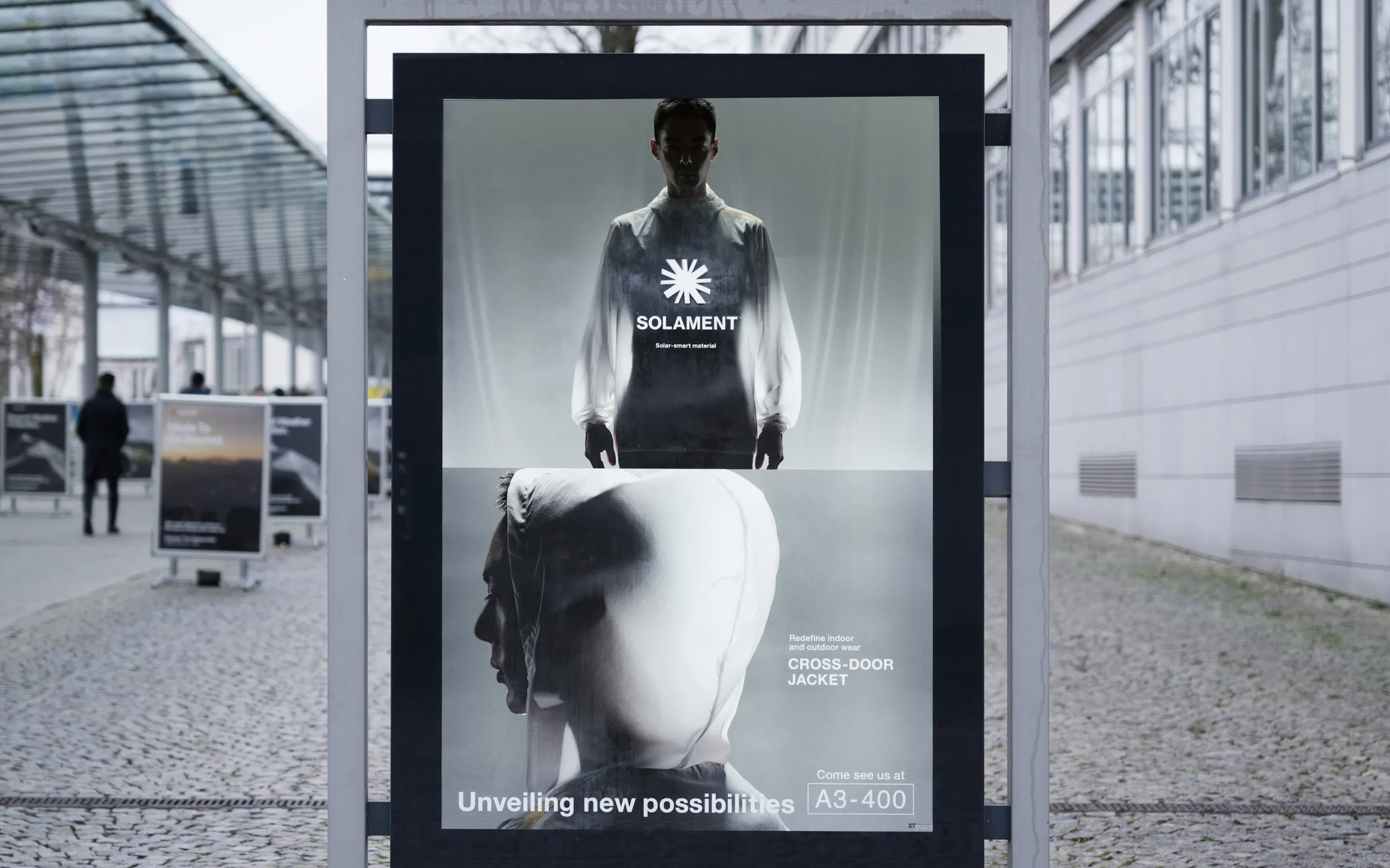 A visual of a large-framed poster featuring the SOLAMENT™ brand’s CROSS-DOOR JACKET installed along the outdoor walkway at ISPO