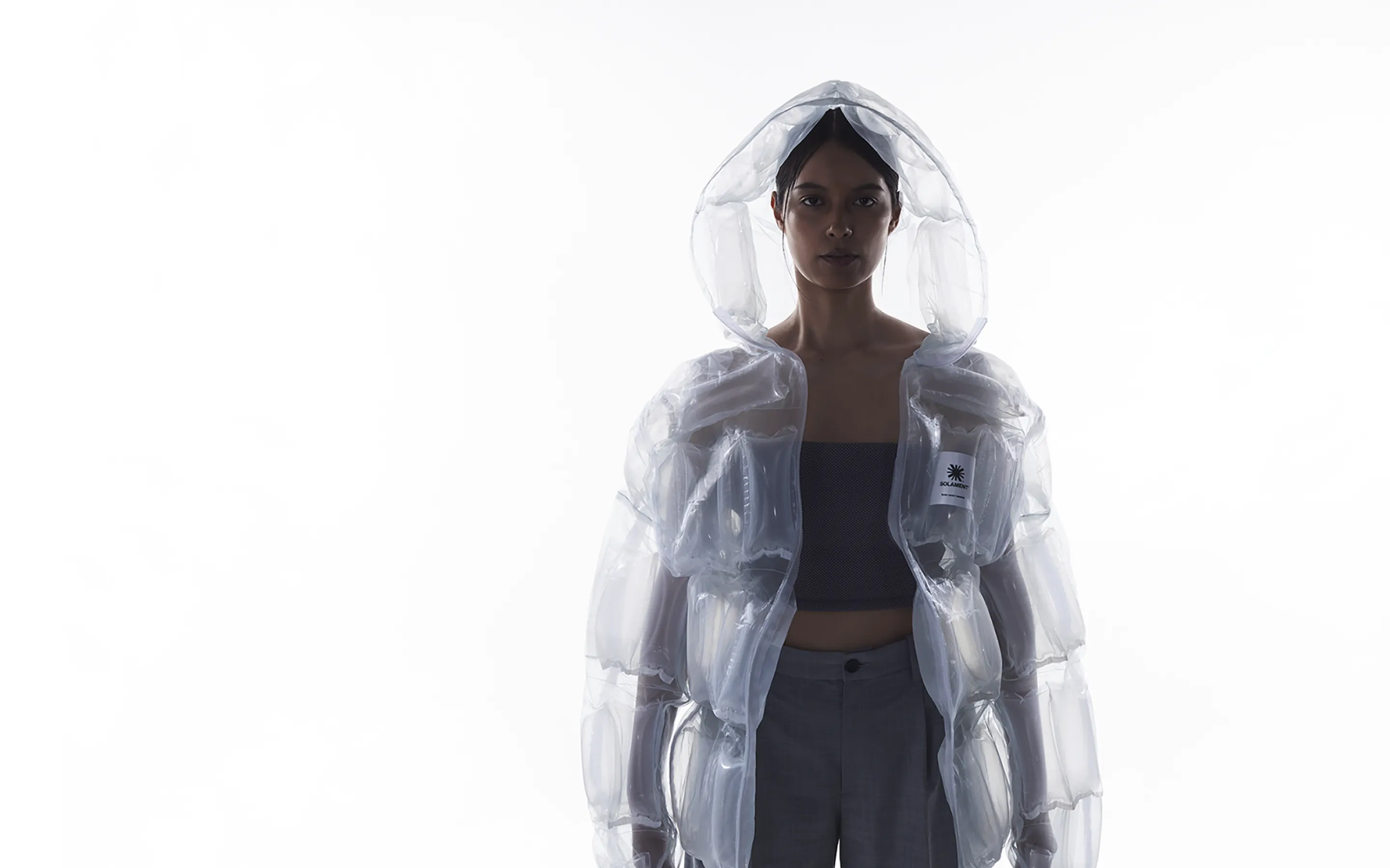 Woman wearing a transparent hooded jacket, DOWN-LESS DOWN JACKET, expressing high functionality through an air-filled structure using minimal resources.