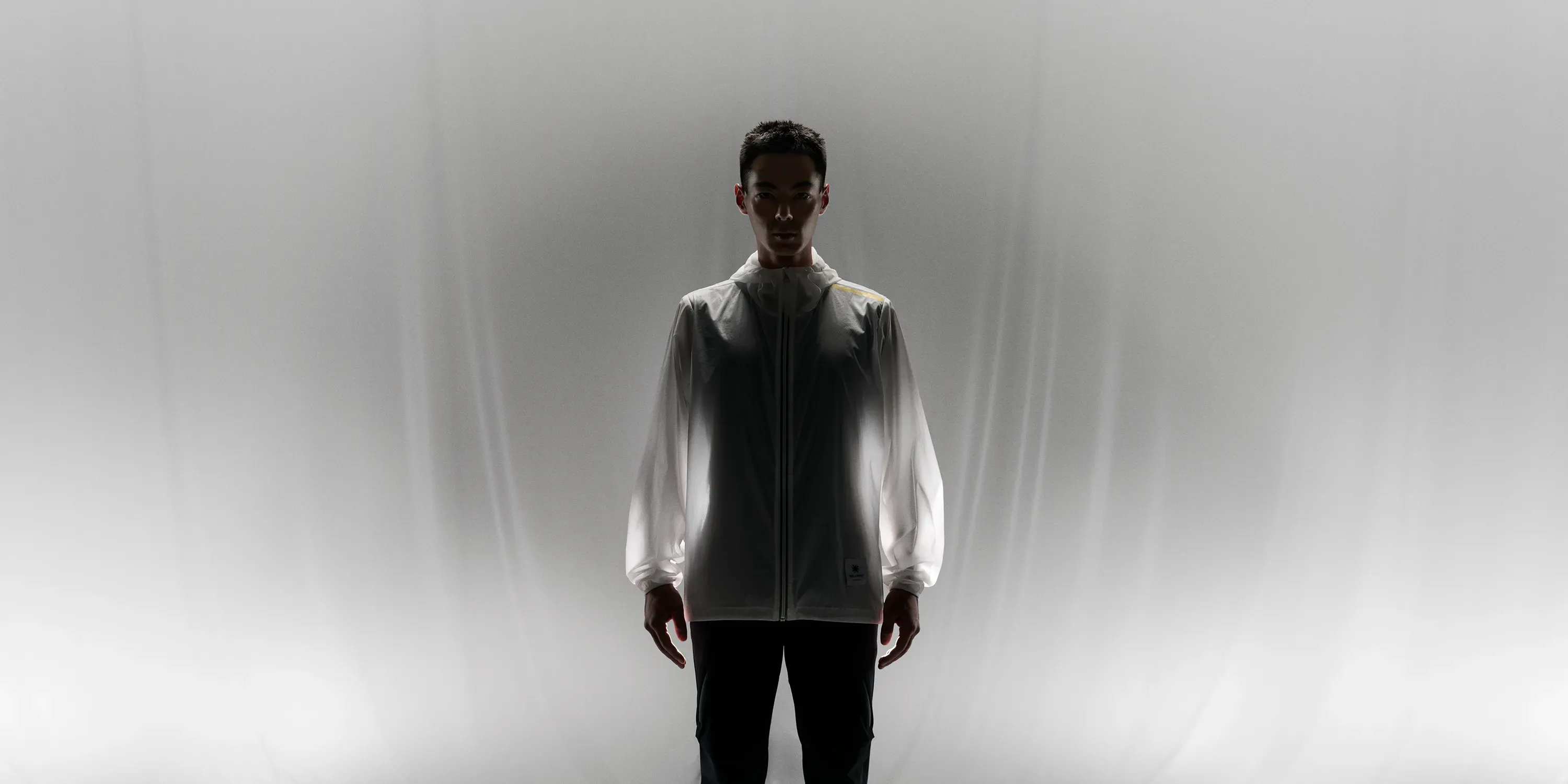Man in strong backlight, with the CROSS-DOOR JACKET glowing white, expressing thin fabric and comfortable heat-generation.