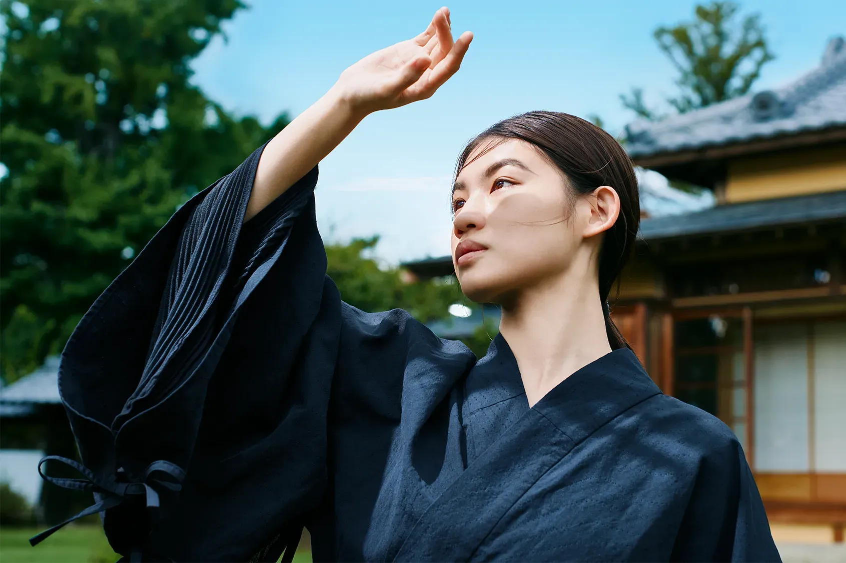 Woman wearing HOPE WEAR for Culture in front of a traditional Japanese house, raising her arms to show the breathable pleated design inside the wide sleeves.