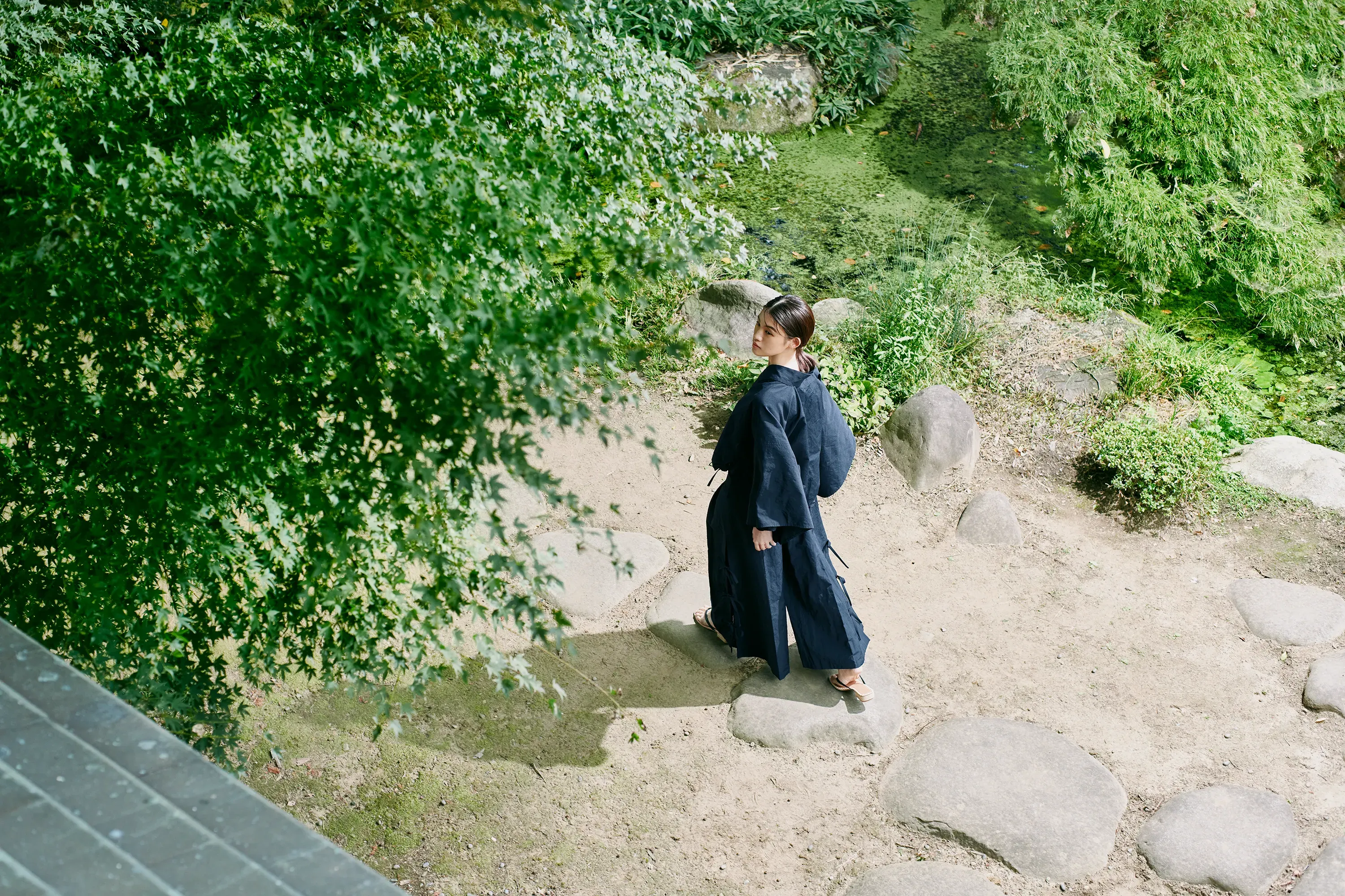 Woman walking in a Japanese garden, wearing a deep navy kimono-inspired setup, HOPE WEAR for Culture, with wide sleeves showing summer comfort.