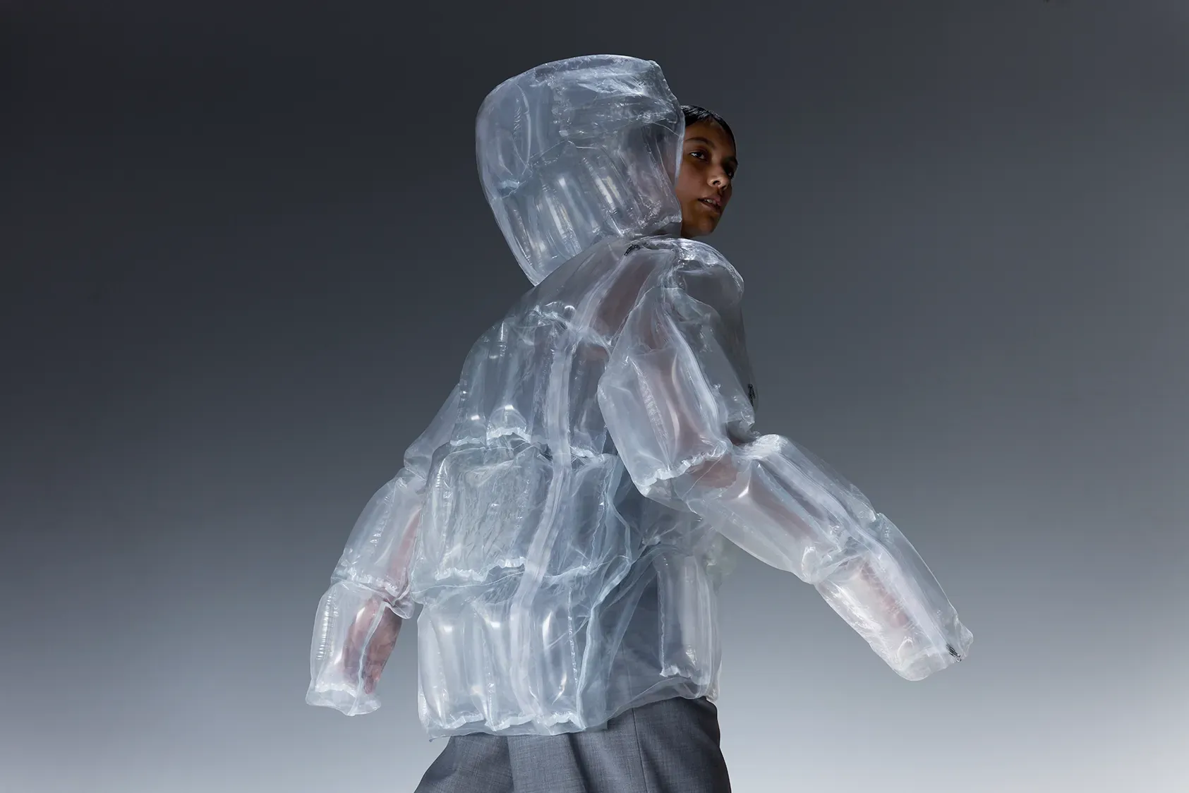 Model looking back in a transparent DOWN-LESS DOWN JACKET, made without down and constructed with air layers that generate heat from sunlight.