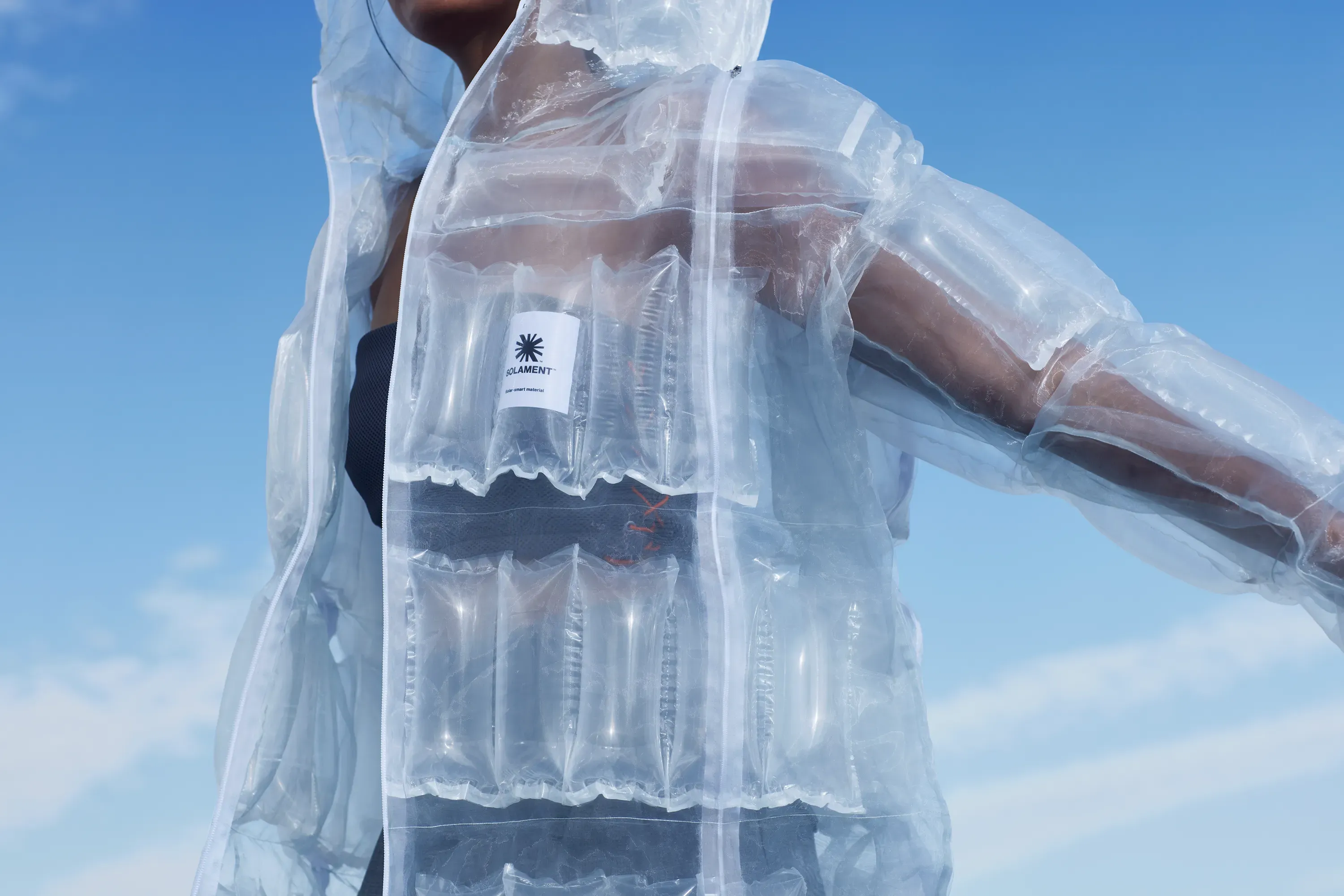 Model in a transparent DOWN-LESS DOWN JACKET under a blue sky, with sunlight heat‑generating air blocks visible through the fabric.