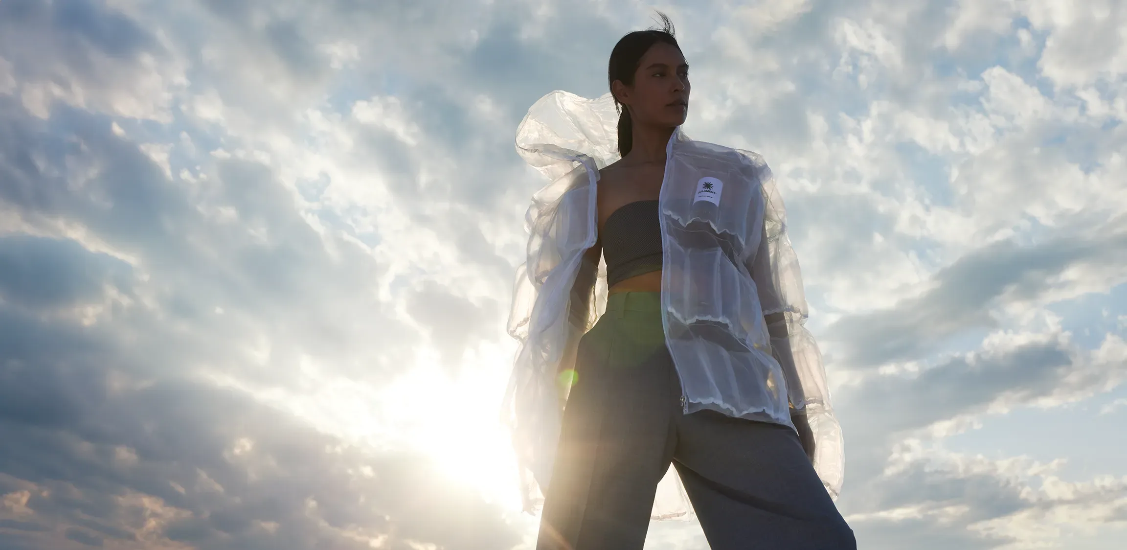 Model standing against strong sunlight and clouds, expressing the functionality of the transparent DOWN-LESS DOWN JACKET.