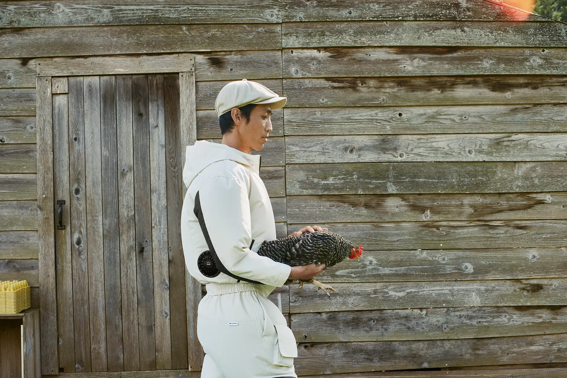 A man holding a chicken, wearing off-white agricultural workwear "HOPE WEAR for Farm Type A," equipped with built-in KUCHOFUKU fans at the waist.