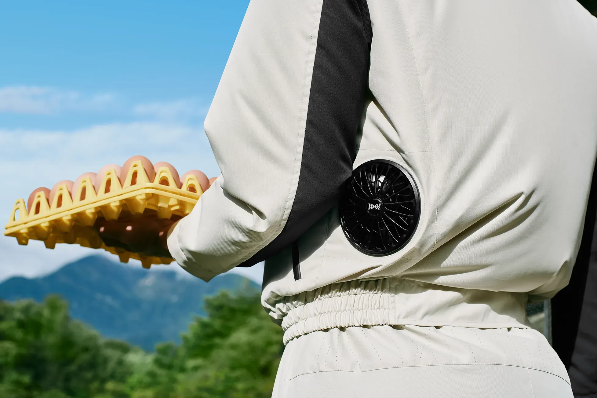 Seen from behind, a person on a farm holds a yellow egg tray, wearing an off-white "HOPE WEAR for Farm Type A" jacket with waist "KUCHOFUKU" fans.