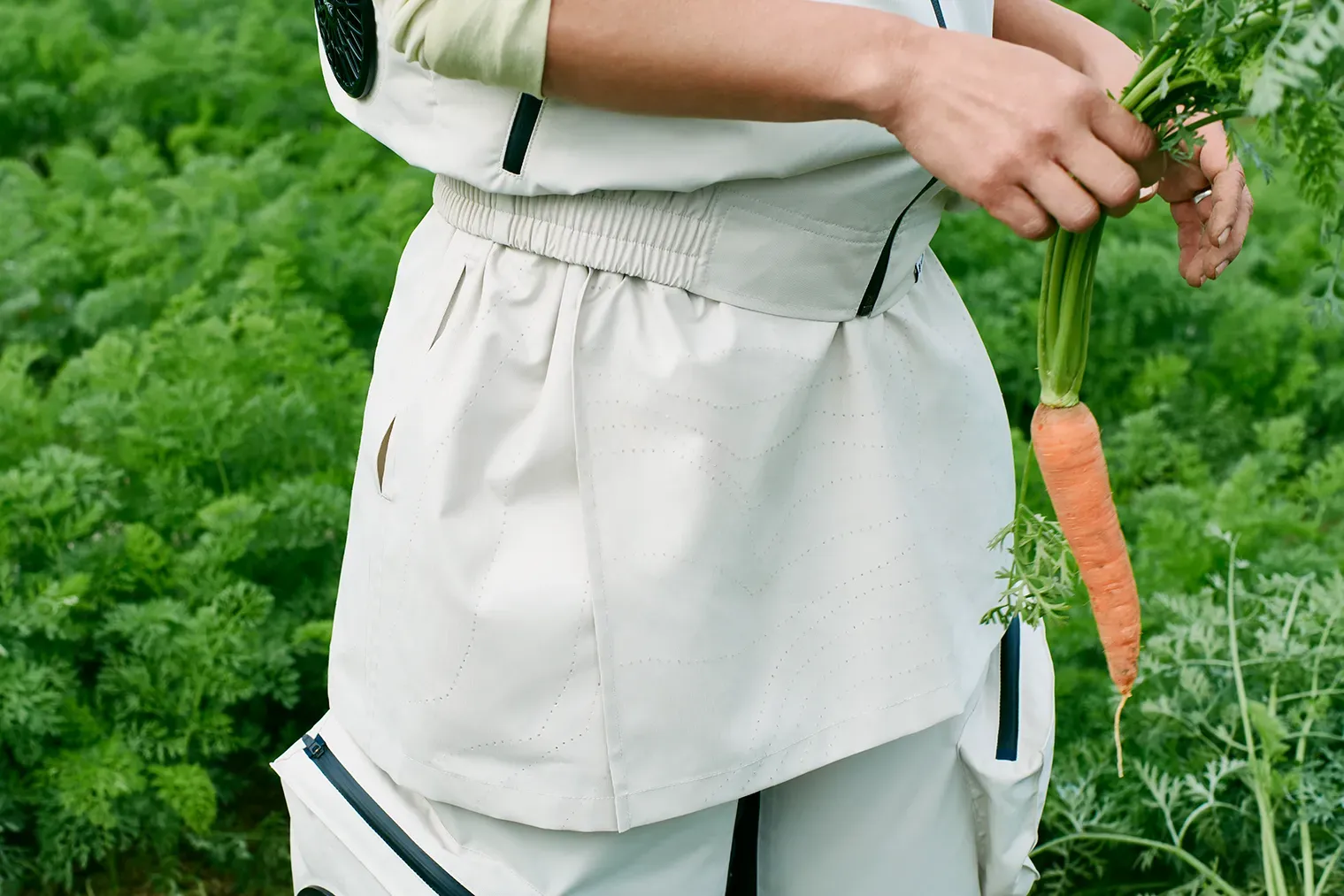 Close-up of a farm worker wearing HOPE WEAR Type B pants with apron and fans at waist, holding fresh carrots in a green field.