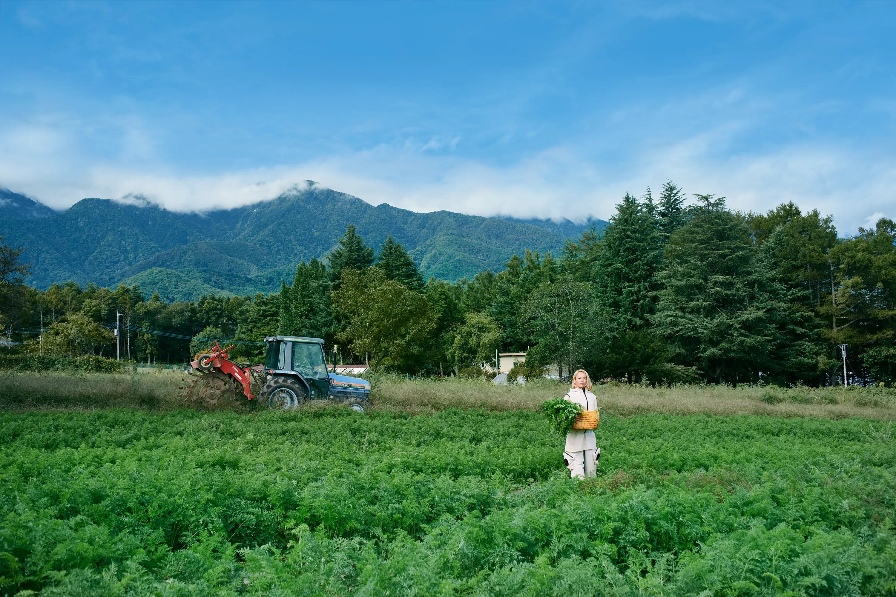 Woman holding a vegetable basket, wearing HOPE WEAR for Farm Type B with farmland behind her and a tractor working in the distance.