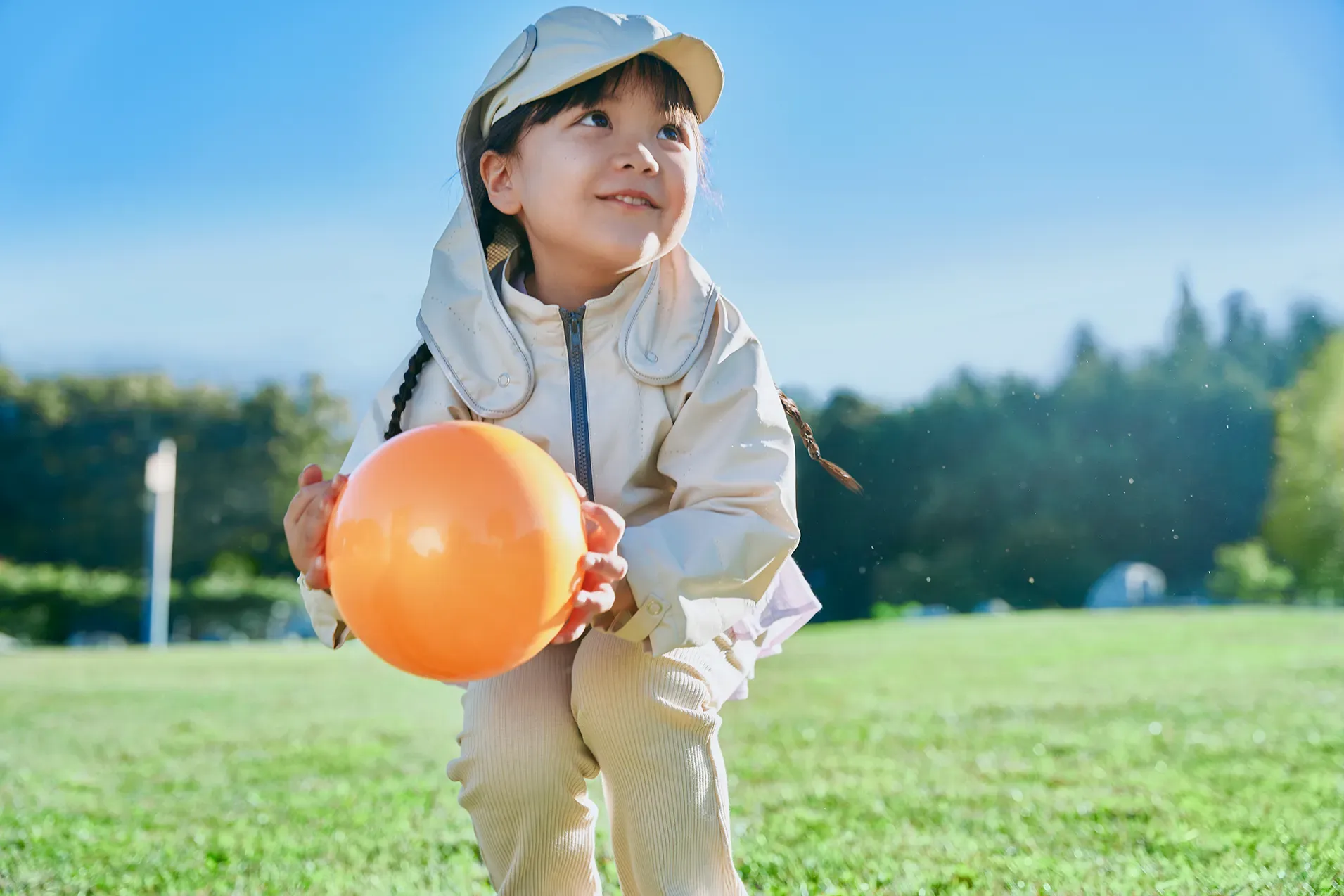 Girl holding an orange ball under a blue sky, wearing a sun-shade hat and a functional jacket, HOPE WEAR for Play.